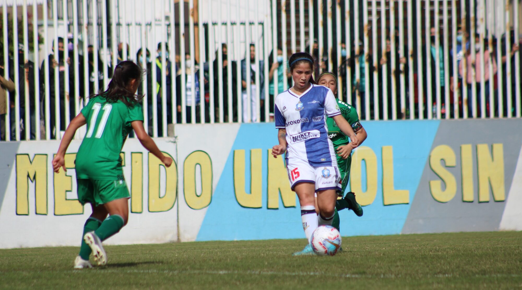 Una jugadora de Antofagasta con la pelota y dos de Deportes Temuco alrededor. Atrás personas viendo el partido por la reja esperando el retorno al estadio